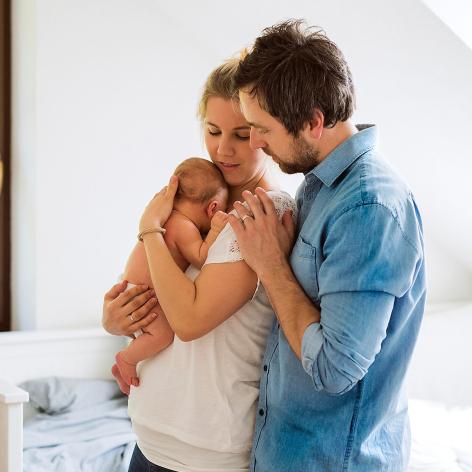 Mutter hält das Baby im Arm, während der Vater liebevoll eine Hand auf ihre Schulter legt