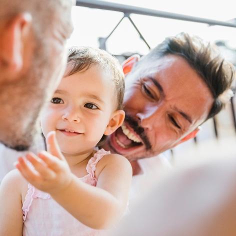 Ein Mann hält ein lachendes Kleinkind von hinten im Arm. Das Kind streckt freudig den Arm zu einem anderen Mann gegenüber aus.